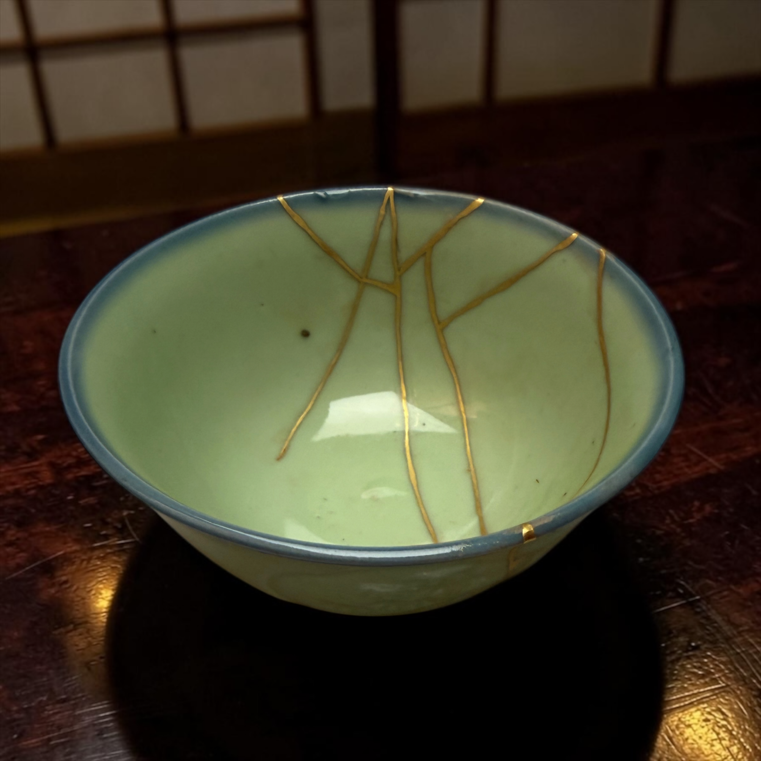 IMG_2563 | Kintsugi by Myriam GREFF Close-up of a light green ceramic bowl with gold decorative lines resting on a dark wooden table edge visible in the frame.