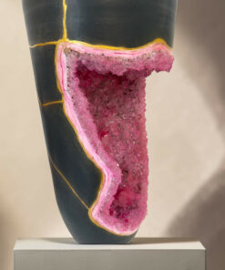 Close-up of a pink crystal geode peeking from a dark green vase with a yellow seam, set on a white pedestal.