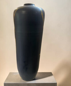 Black ceramic vase with gold kintsugi cracks on a light pedestal against a neutral wall.