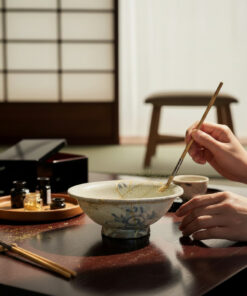Hands stir a ceramic bowl with a wooden chopstick on a dark wooden table; a tray with small bottles sits to the left.