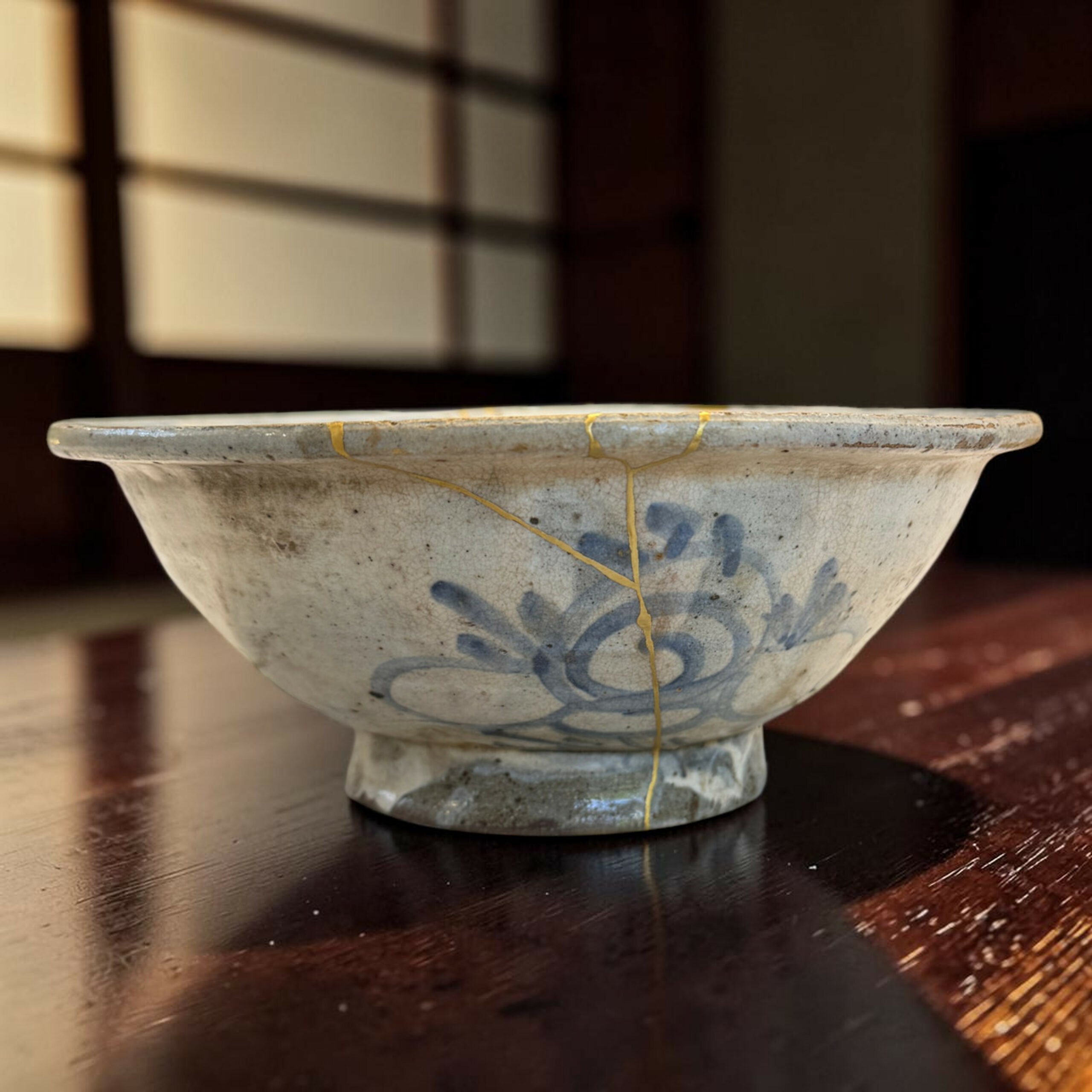 IMG_2969-Photoroom | Kintsugi by Myriam GREFF Close-up of a ceramic bowl with blue floral glaze and gold-toned cracks, resting on a dark wooden table.