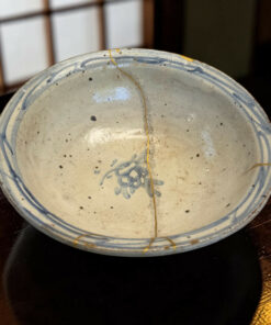Close-up of a ceramic plate with a blue patterned rim and a gold crack running across its light glaze on a dark wooden table.