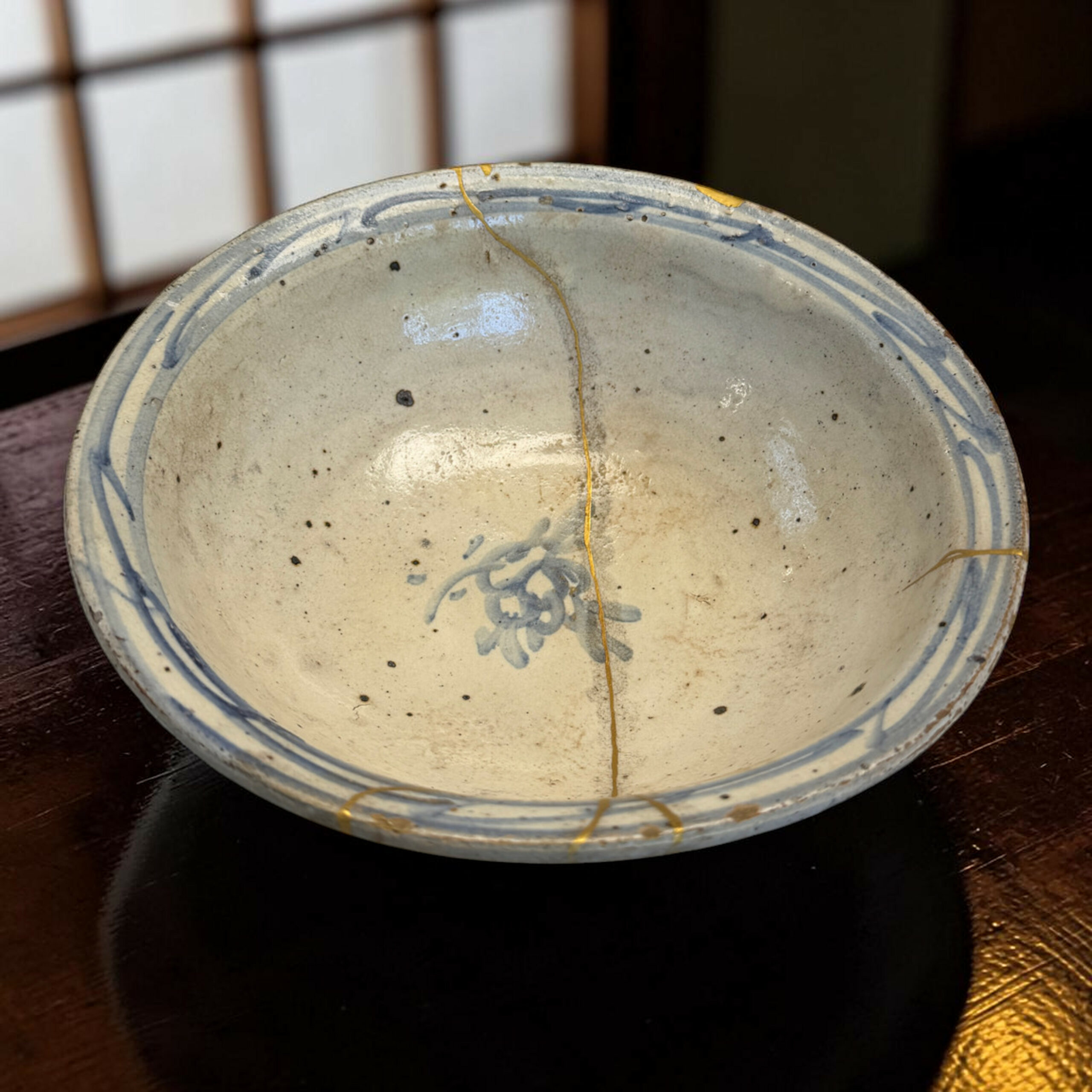 IMG_2970-Photoroom | Kintsugi by Myriam GREFF Close-up of a ceramic plate with a blue patterned rim and a gold crack running across its light glaze on a dark wooden table.