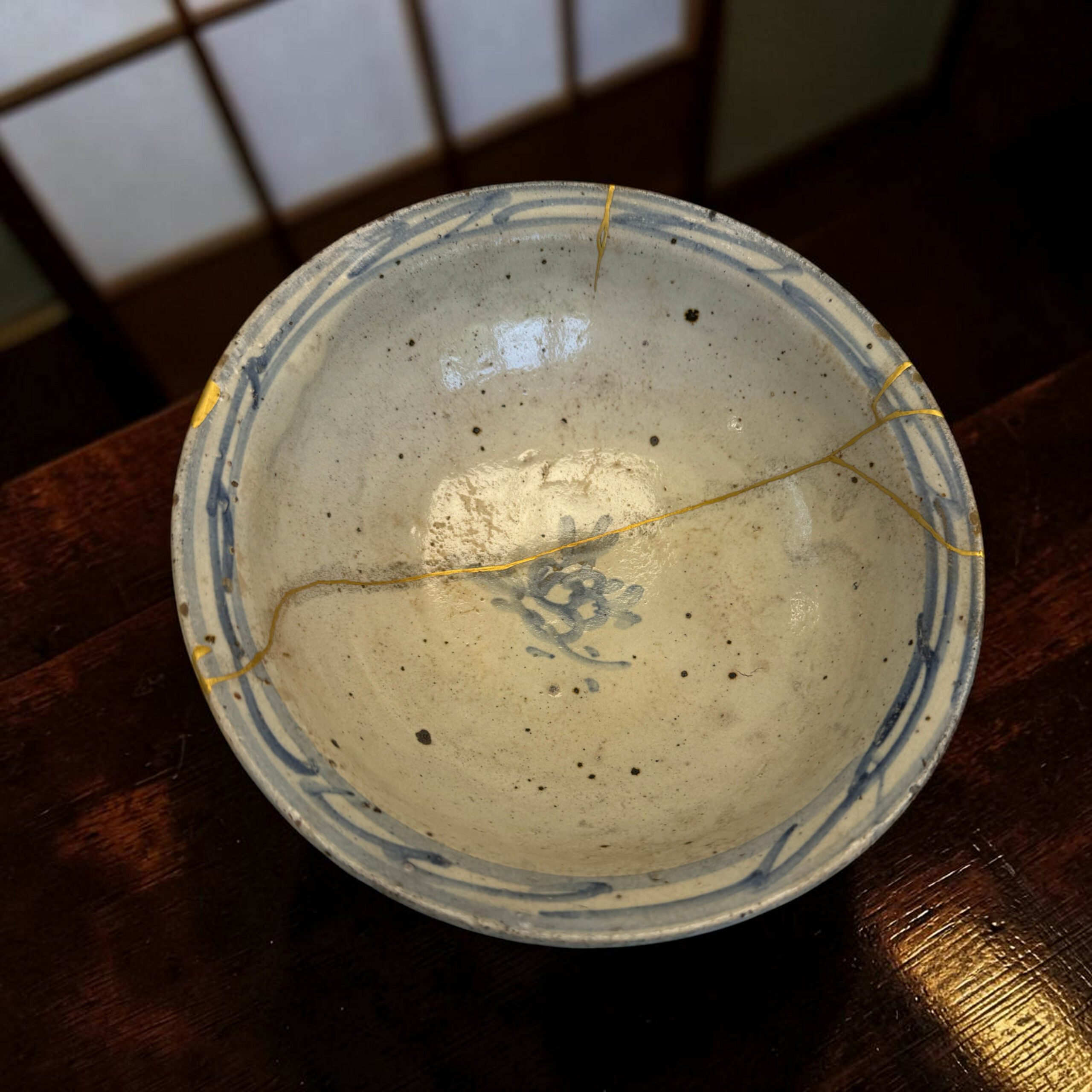 IMG_2972-Photoroom | Kintsugi by Myriam GREFF Close-up of a speckled ceramic plate with a blue rim placed on a dark wooden table, with a thin gold string resting on it.