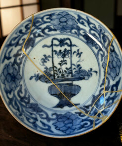 Blue-and-white porcelain plate with a decorative pattern, showing gold repair seams (kintsugi) and a tree motif, on a wooden surface
