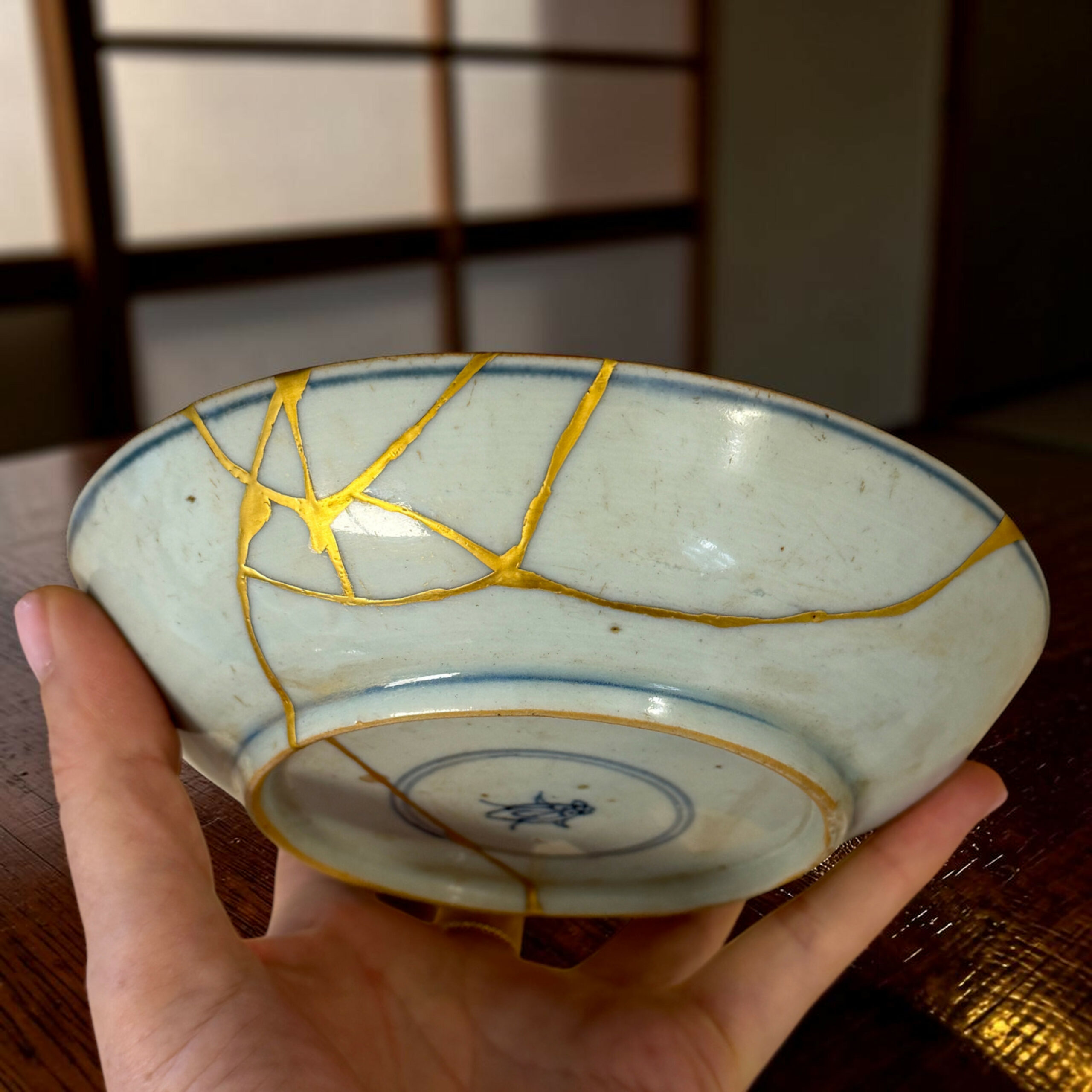 IMG_3117-Photoroom | Kintsugi by Myriam GREFF Hands cradle a pale blue ceramic plate with gold crackle lines and blue trim, resting on a wooden surface