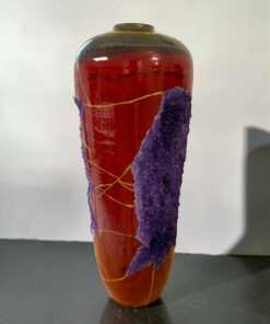Close-up of a tall red glass vase wrapped with purple fuzzy material and yellow string, on a dark table.