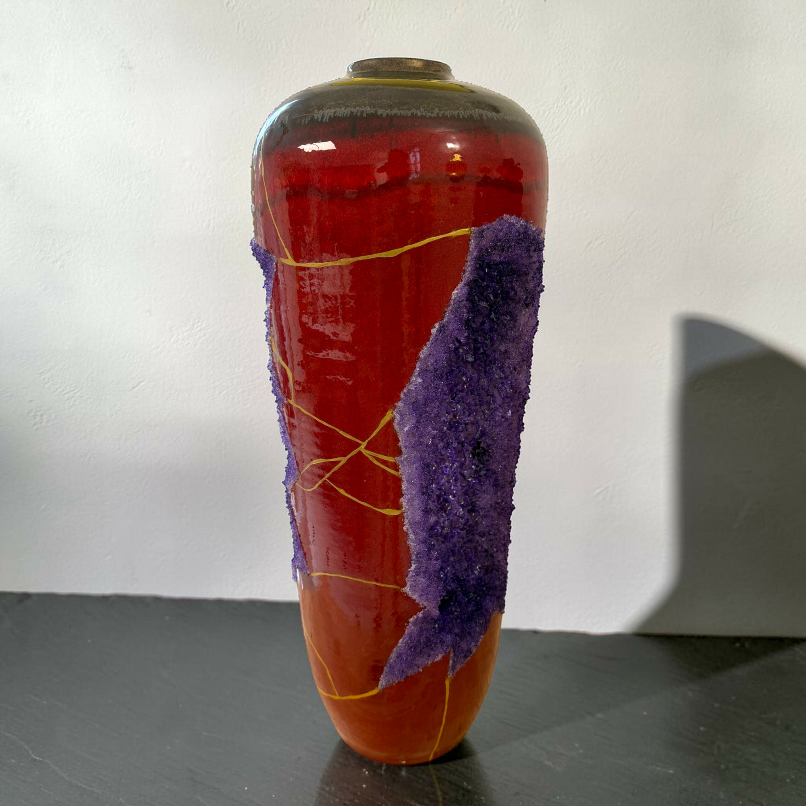 IMG_3238-Photoroom | Kintsugi by Myriam GREFF Close-up of a tall red glass vase wrapped with purple fuzzy material and yellow string, on a dark table.