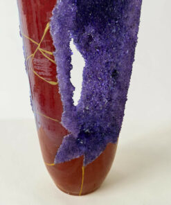 Close-up of a red ceramic vase wrapped with purple glittery material and yellow string against a pale background.