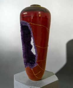 Red ceramic vase with purple crystallized deposits and a yellow line, displayed on a gray stepped pedestal.