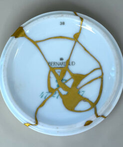 Close-up of a pale blue ceramic plate with tangled gold ribbon laid across its surface; the number 38 is visible near the top edge.