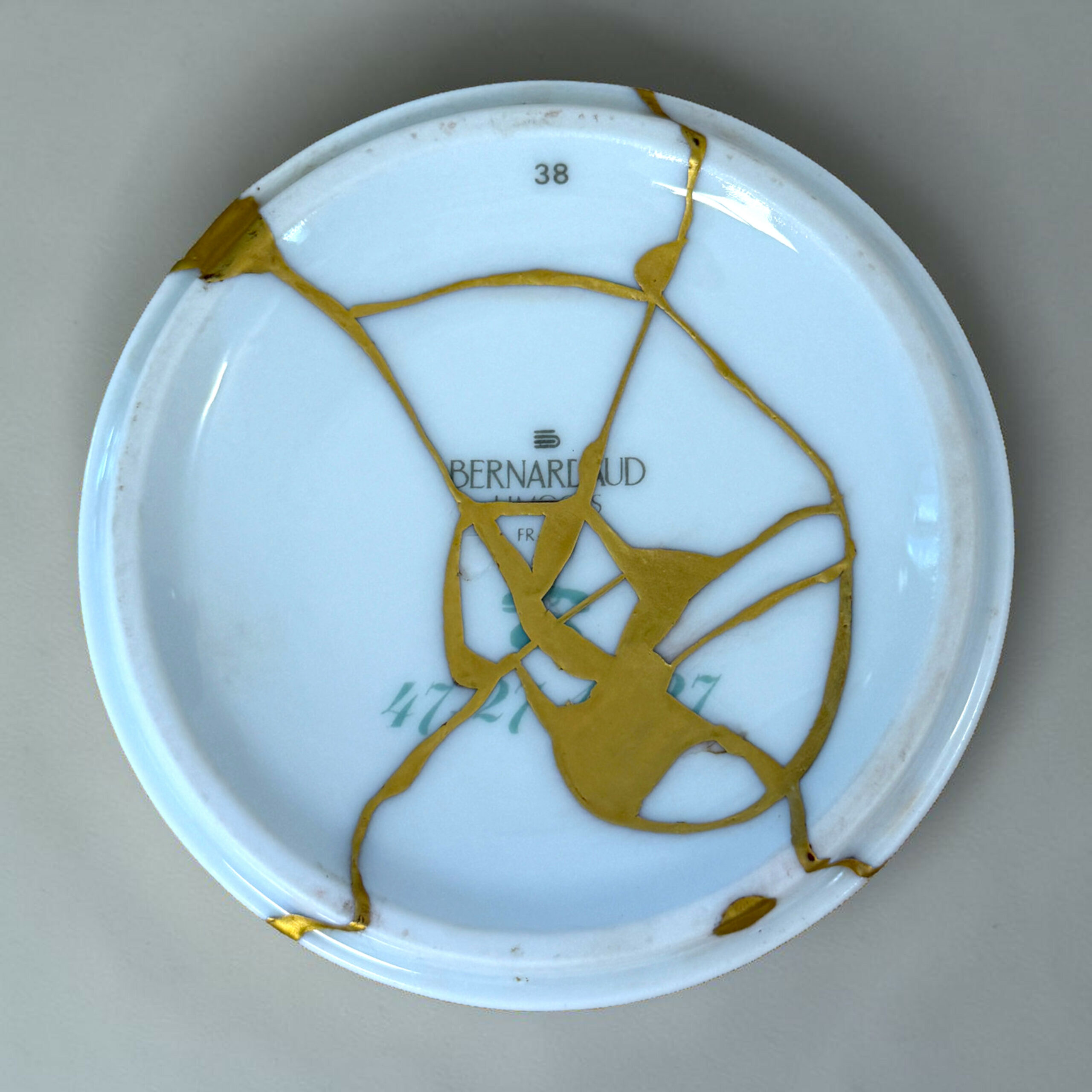 ducasse 1 | Kintsugi by Myriam GREFF Close-up of a pale blue ceramic plate with tangled gold ribbon laid across its surface; the number 38 is visible near the top edge.