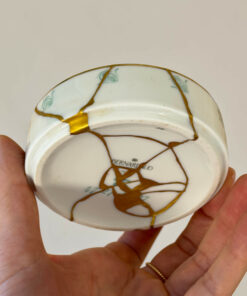 Hands hold a white porcelain plate with prominent gold-filled cracks (kintsugi) across its surface.