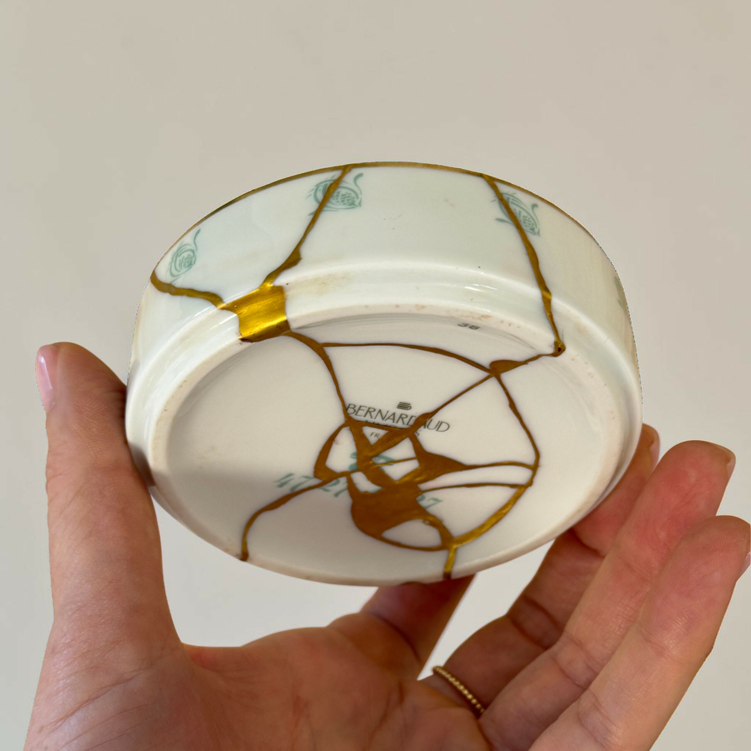 ducasse 2 | Kintsugi by Myriam GREFF Hands hold a white porcelain plate with prominent gold-filled cracks (kintsugi) across its surface.