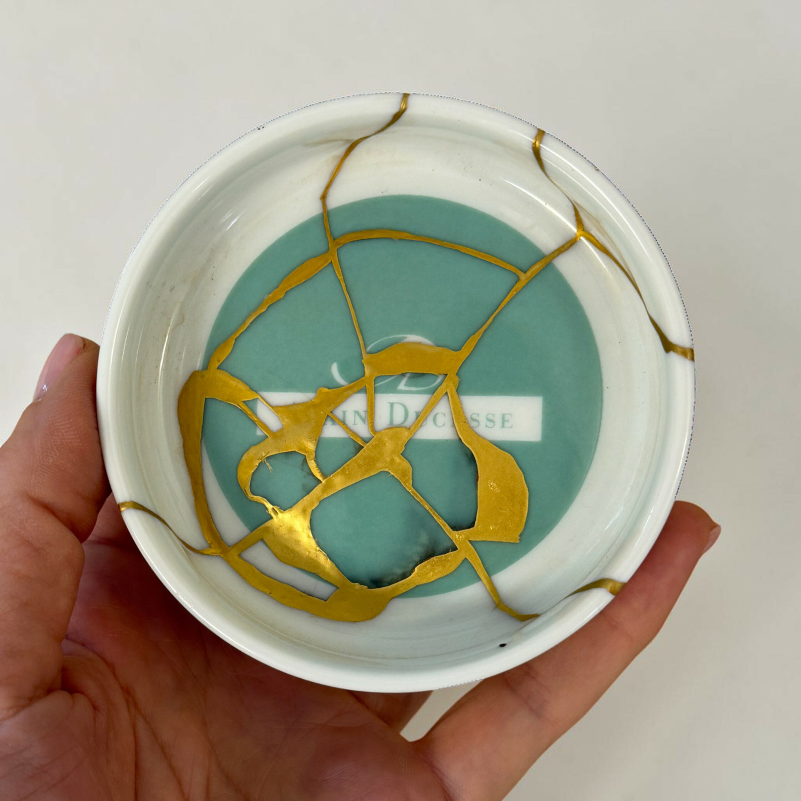 ducasse 3 | Kintsugi by Myriam GREFF Close-up of a ceramic plate with teal center and gold crackle pattern, held in a person's hands